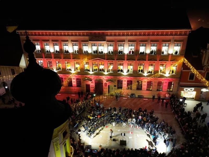 Pogled iznad na Božićni koncert na prozorima Gimnazije Matije Antuna Reljkovića u Vinkovcima, osvijetljena zgrada i publika na trgu